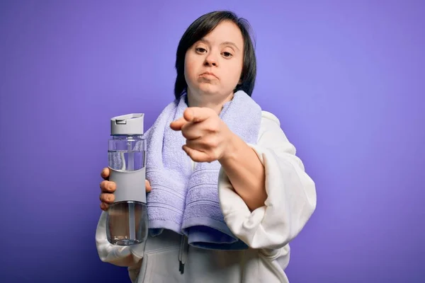 Young down syndrome fitness woman training at gym holding water bottle and sport towel pointing with finger to the camera and to you, hand sign, positive and confident gesture from the front