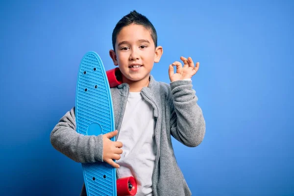 小男孩滑板手抱着现代滑板在蓝色孤立的背景上 用手指做着确定的手势 很好的符号 — 图库照片