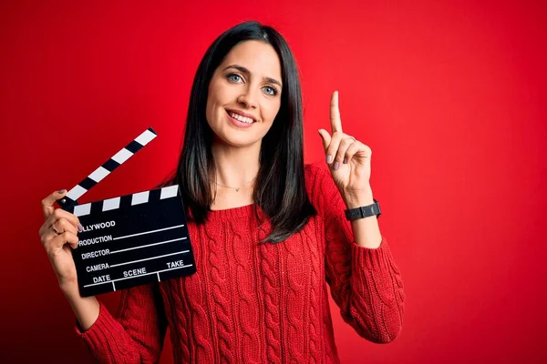 Young director woman with blue eyes making movie holding clapboard over red background surprised with an idea or question pointing finger with happy face, number one