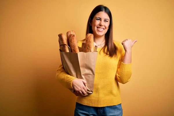 年轻美丽的女人 提着一袋黄色背景的健康的新鲜面包 手背朝后 竖起大拇指 带着自信的微笑 — 图库照片