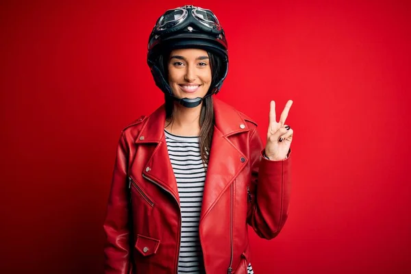 若い美しいブルネットのオートバイのヘルメットと赤いジャケットを身に着けているオートバイの女性は 勝利のサインを行うカメラで顔をウィンクして幸せな顔で笑っています 第二番 — ストック写真