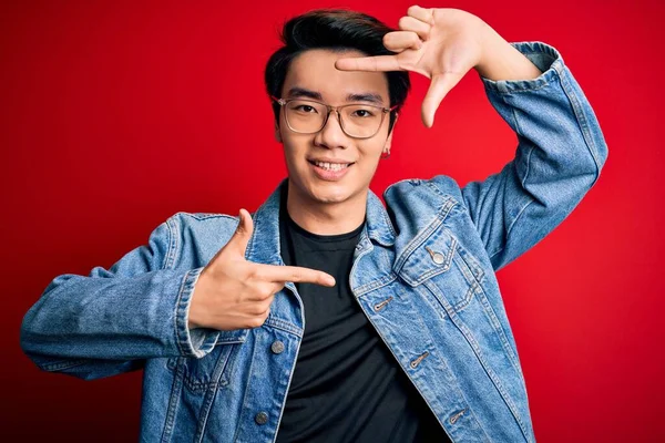 Young handsome chinese man wearing denim jacket and glasses over red background smiling making frame with hands and fingers with happy face. Creativity and photography concept.