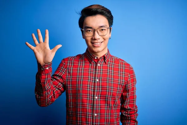 若いですハンサムな中国人男性身に着けていますカジュアルシャツと眼鏡上の青の背景表示と指差す指番号5ながら笑顔自信と幸せ — ストック写真