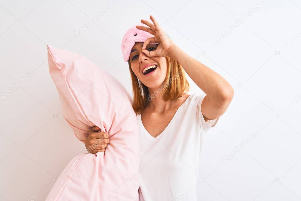 Beautiful redhead woman wearing sleeping mask cuddling pink pillow over isolated background with happy face smiling doing ok sign with hand on eye looking through fingers