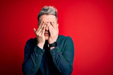 Kırmızı izole edilmiş arka plan üzerinde zarif yeşil tişört giyen genç, yakışıklı ve modern adam yorgunluk ve baş ağrısı için gözlerini ovuşturuyor, uykulu ve yorgun yüz ifadesi takınıyor. Görüş sorunu