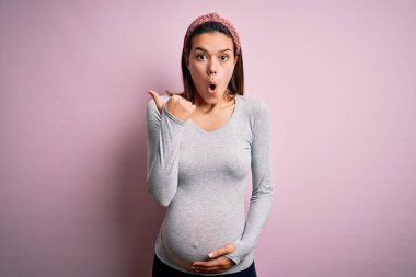 Genç, güzel bir kız hamile, pembe arka planda bebek bekliyor. El parmağı yana dönük, ağzı açık, şaşırmış bir ifade..