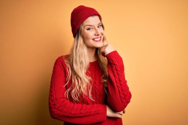 Beyaz arka planda günlük kazak ve yün şapka giyen genç güzel sarışın kadın çapraz kollarla gülümseyen ve elleri çenesinde yükselen kameraya kendinden emin bir şekilde bakıyor. Olumlu düşünüyorum..