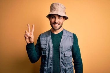 Tatilde sakallı yakışıklı turist sarı arka planda kaşif şapkası takıyor ve kameraya bakıp zafer işareti yapıyor. İki numara..