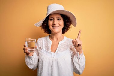 Orta yaşlı kıvırcık kadın tatilde izole edilmiş sarı arka planda su içiyor. Bir fikir ya da soru işareti ile şaşırıyor.