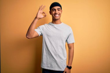 Yakışıklı Afro-Amerikan sporcu sarı arka planda spor giysisi giyiyor gülümsüyor ve el ve parmaklarıyla tamam işareti yapıyor. Başarılı ifade.