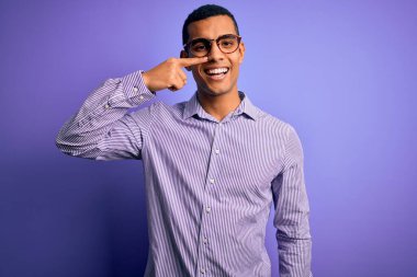 Yakışıklı Afro-Amerikalı, çizgili gömlek ve gözlük takmış, mor arka planda, eli ve burnu işaret eden, neşeli gülümseyen. Güzellik konsepti