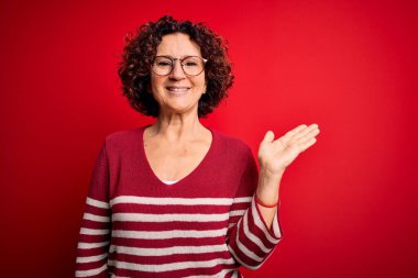 Orta yaşlı, kıvırcık saçlı, sıradan çizgili süveter giyen, kırmızı arka planda gülümseyen, eli kameraya bakan güzel bir kadın..