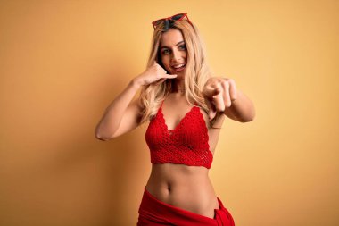 Sarışın, izole edilmiş sarı arka planda bikinili genç ve güzel bir kadın telefonda konuşurken gülümsüyor ve seni işaret ediyor. Ara beni..