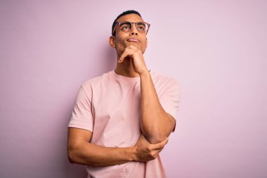 Yakışıklı, Afro-Amerikalı bir adam pembe arka plan üzerinde günlük tişört ve gözlük takıyor ve elleri çenesinde soru sormayı düşünüyor. Düşünceli bir ifade. Gülen ve düşünceli bir yüz. Şüphe kavramı.