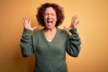 Orta yaşlı, kıvırcık saçlı güzel bir kadın izole edilmiş sarı arka plan üzerine gündelik süveter giyip kollarını kaldırmış ve gözleri kapalı heyecanla haykırarak çılgın ve çılgın bir başarıyı kutluyor. Kazanan konsept