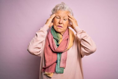 Son sınıftaki güzel, gri saçlı kadın pembe arka planda günlük süveter ve eşarp giyiyor. Ağrı ve migrenden dolayı çaresiz ve stresli. Ellerini başının üstüne koy..