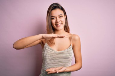 Genç, güzel esmer kız izole edilmiş pembe arka plan jestlerinin üzerine sıradan çizgili tişört giyiyor. Ellerinde büyük ve büyük beden işareti, ölçü işareti var. Kameraya bakıp gülümsüyor. Ölçüm konsepti.