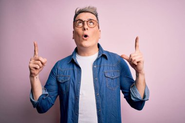 Pembe izole arka planda gözlük ve kot ceket giyen genç yakışıklı modern adam şaşırmış ve şaşırmış bir şekilde yukarı bakıyor ve parmaklarıyla işaret ediyor ve kollarını kaldırıyor..