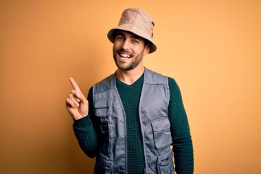 Tatilde sakallı yakışıklı turist sarı arka planda kaşif şapkası takıyor, yüzünde kocaman bir gülümsemeyle kameraya el ile işaret ediyor..
