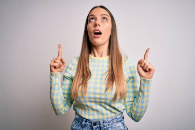 Genç, güzel, sarışın, mavi gözlü, izole edilmiş arka planda duran bir kadın. Şaşırmış ve şaşırmış bir şekilde yukarı bakıyor ve parmaklarıyla işaret ediyor ve kollarını kaldırıyor..