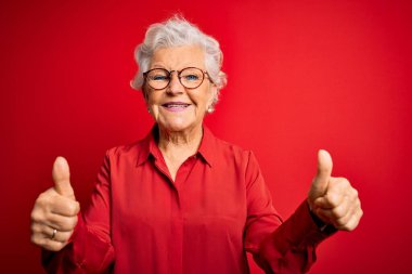 Kıdemli, gri saçlı, günlük gömlek ve gözlük takan, kırmızı arka planda olumlu jestler yapan, gülümseyen ve başarı için mutlu bir kadın. Kazanan jest.