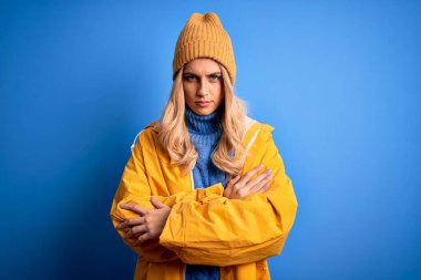 Mavi arka planda yağmurlu hava için yağmurluk giyen genç ve güzel sarışın kadın şüpheci ve gergin, yüzünde çapraz kollu tasvip etmeyen bir ifade var. Negatif kişi.