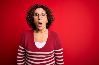 Orta yaşlı güzel kıvırcık saçlı kadın kırmızı arka planda sıradan çizgili süveter giyiyor. Şaşırtıcı bir ifade, korku ve heyecan dolu bir yüz ifadesi var..