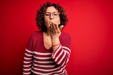 Orta yaşlı güzel kıvırcık saçlı kadın kırmızı arka planda sıradan çizgili süveter giyiyor kameraya bakıyor ve havada elle öpüşüp güzel ve seksi oluyor. Aşk ifadesi.