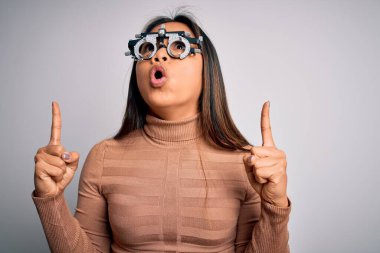 Genç Asyalı optik kız beyaz arka planda optometri gözlüğü kullanarak görme yetisini kontrol ediyor. Şaşırmış ve şaşırmış bir şekilde yukarı bakıyor ve parmaklarıyla işaret ediyor ve kollarını kaldırıyor..