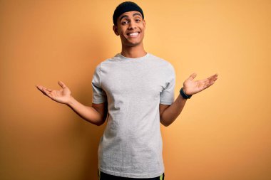 Yakışıklı Afro-Amerikan sporcusu sarı arka planda spor giysisi giyiyor. Gülümsüyor, iki elini de açık gösteriyor. Karşılaştırma ve reklam karşılaştırması yapıyor ve dengeyi sağlıyor.