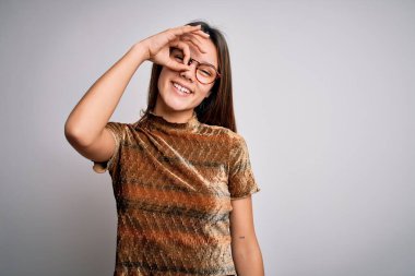 Güzel, esmer, günlük tişört ve gözlük takan genç bir kız izole edilmiş beyaz arka planda el hareketleriyle gülümsüyor ve mutlu yüzüyle parmaklarının arasından bakıyor..