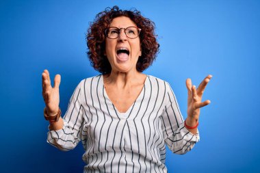 Orta yaşlı, güzel kıvırcık saçlı, sıradan çizgili tişörtlü bir kadın izole arka planda çılgınca bağırıyor ve agresif bir ifadeyle bağırıyor ve kollarını kaldırıyor. Hayal kırıklığı konsepti.