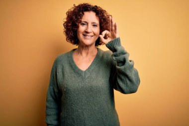 Orta yaşlı, kıvırcık saçlı güzel bir kadın izole edilmiş sarı arka plan üzerine günlük süveter giyip el ve parmaklarıyla 