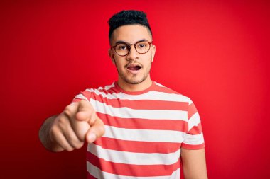 Genç, çizgili tişört ve gözlük takan yakışıklı bir adam izole edilmiş kırmızı arka planda kameraya kızgın ve sinirli olduğunu gösteriyor.