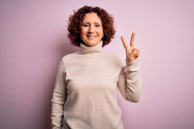 Orta yaşlı güzel kıvırcık saçlı kadın pembe arka planda balıkçı yaka kazak giyiyor ve parmaklarıyla iki numarayı gösterirken kendinden emin ve mutlu gülümsüyor..