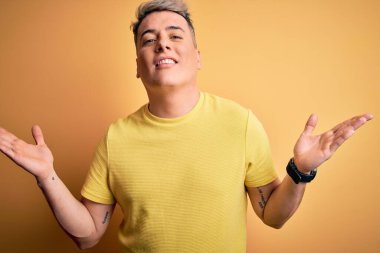 Sarı izole edilmiş arka plan üzerine sarı tişört giyen yakışıklı ve kafası karışmış, kolları ve elleri havada genç bir adam. Şüphe kavramı.