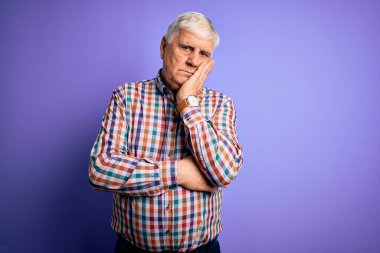 Kıdemli yakışıklı, beyaz gömlekli, yalıtılmış, mor arka planda oturan, bunalımdan sıkılmış ve yorgun görünen, çapraz kollu yaşlı bir adam..