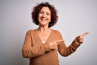 Orta yaşlı güzel kıvırcık saçlı kadın izole edilmiş beyaz arka planda günlük süveter giyiyor gülümsüyor ve kameraya bakıyor iki eliyle ve parmaklarıyla yandan işaret ediyor..