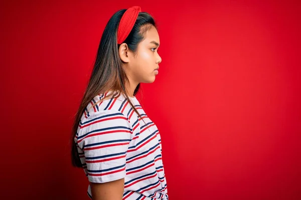 年轻美丽的亚洲女孩穿着宽松的条纹T恤 披着孤零零的红色背景 向侧面看去 以自然的面容和自信的微笑放松自己的姿态 — 图库照片