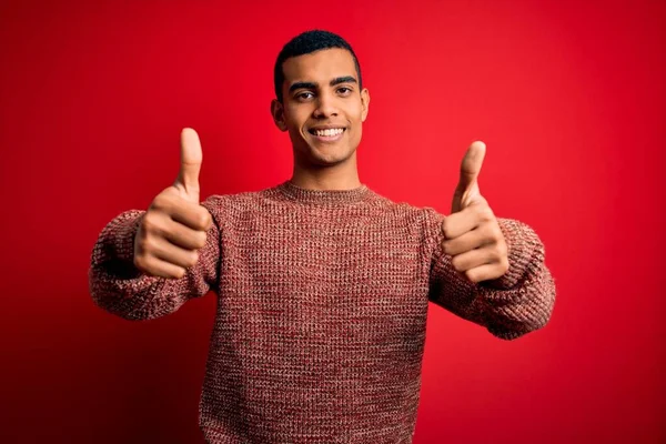 赤い背景の上にカジュアルなセーターを着た若いハンサムなアフリカ系アメリカ人男性が手で積極的なジェスチャーを行うことを承認し 親指を上げて笑顔と成功のために幸せ 勝者のジェスチャー — ストック写真