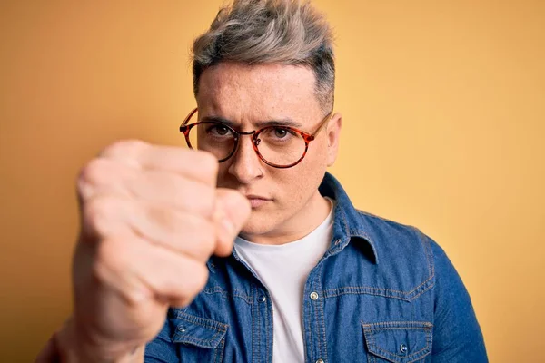 Close up of young handsome modern man wearing glasses and denim jacket over yellow background annoyed and frustrated shouting with anger, crazy and yelling with raised hand, anger concept