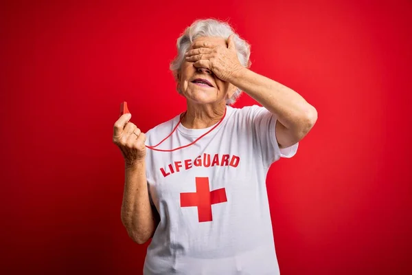年长美丽的灰白头发的救生员女人穿着印有红十字会标志的T恤衫 她面带微笑地吹口哨 用手捂住眼睛笑以表示惊讶 盲目概念 — 图库照片