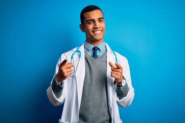 青の背景の上にコートと聴診器を身に着けているハンサムなアフリカ系アメリカ人医師は 幸せと面白い顔を持つカメラに指を指しています 良いエネルギーと振動 — ストック写真
