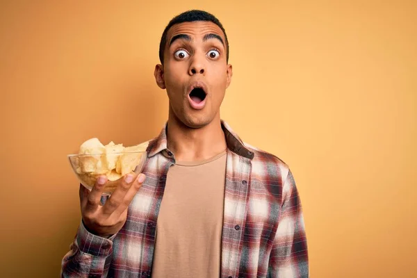 年轻英俊的美国非洲男子 背景是黄色的 手里拿着一碗薯片 吓着了一大跳 脸上带着惊讶的表情 害怕又兴奋 — 图库照片