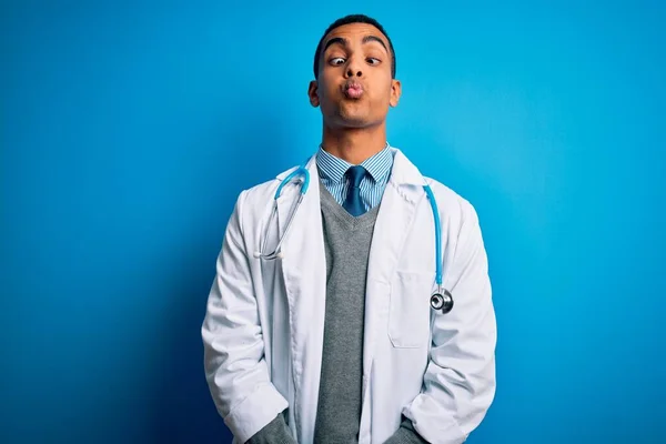 Handsome african american doctor man wearing coat and stethoscope over blue background making fish face with lips, crazy and comical gesture. Funny expression.