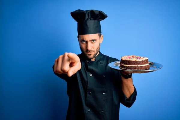 年轻英俊的炊事员 留着胡子 身穿制服 头戴礼帽 手里拿着蛋糕 手指指向相机和你 正面做出积极而自信的手势 — 图库照片