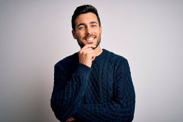 Young handsome man with beard wearing casual sweater standing over white background looking confident at the camera with smile with crossed arms and hand raised on chin. Thinking positive.