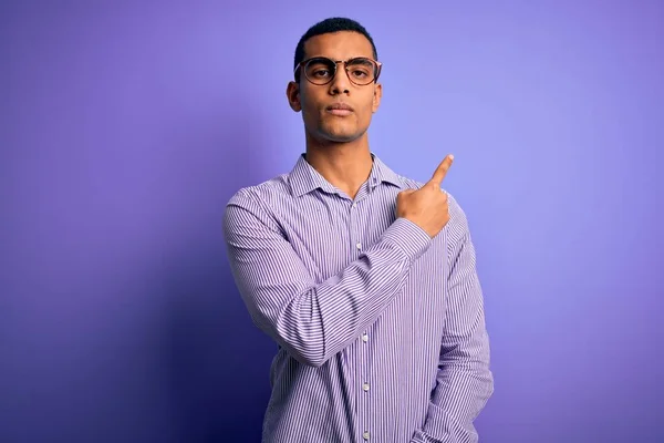 一位英俊的非洲裔美国人 身穿条纹衬衫 眼镜戴在紫色背景上 手指头指向广告的侧面 严肃而沉着 — 图库照片