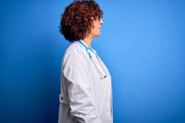 Middle age curly hair doctor woman wearing coat and stethoscope over blue background looking to side, relax profile pose with natural face with confident smile.