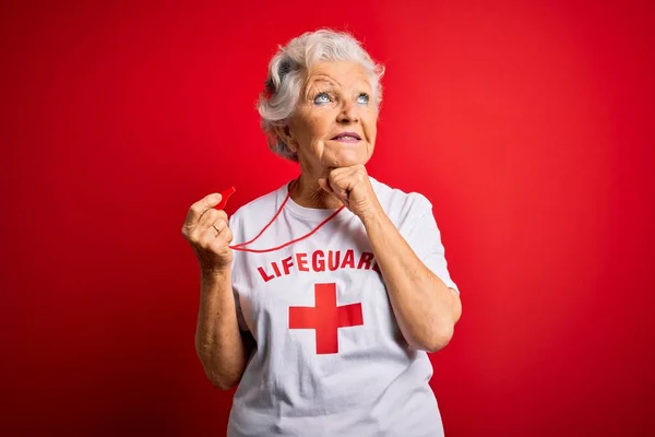 シニア美しい白髪のライフガードの女性は 質問について考える顎の手で笛を使用して赤い十字とTシャツを着て 集中的な表現 笑顔と思慮深い顔 疑いの概念 — ストック写真
